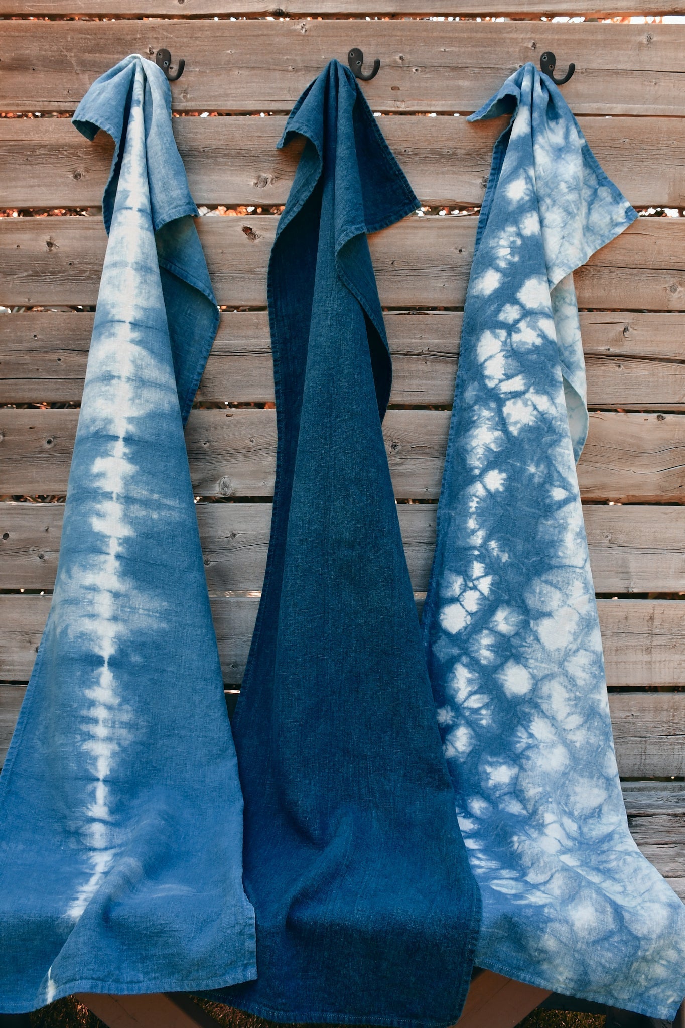 Three indigo-dyed table runners are shown, hung by hooks on a backdrop of weathered barn board.