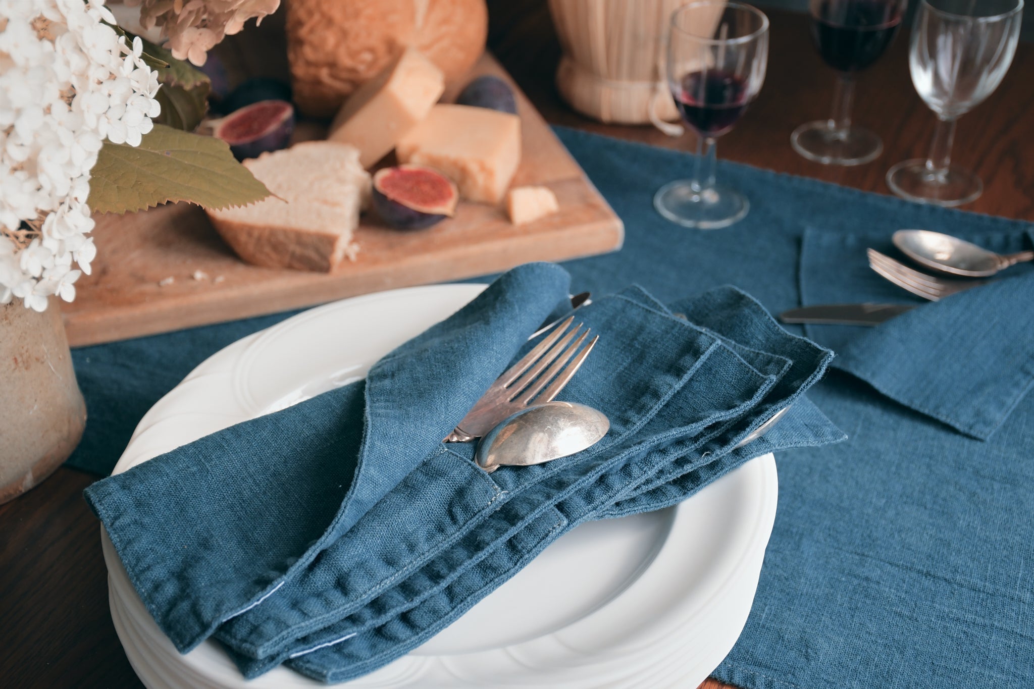 Silverware is tucked into a crafted cutlery wrap, placed on a stack of plates that sits on a warmly set table. a board of cheeses, bread and figs is seen in the background along with wine-filled stemware and some flowers. 