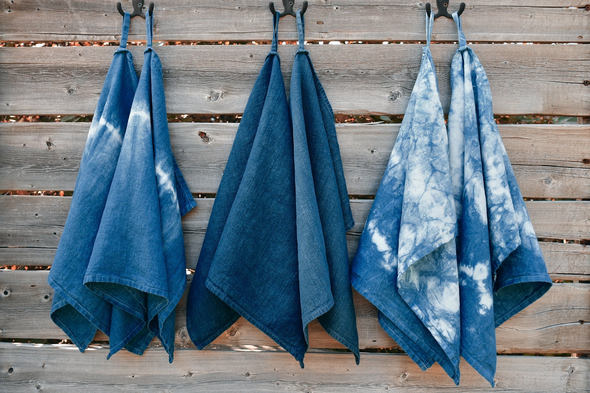 six indigo-dyed tea towels are pictured, hanging from hooks on a weathered wood backdrop